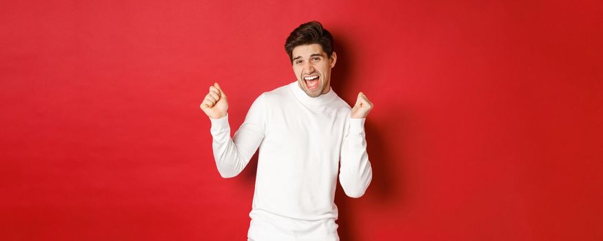 Portrait of handsome man enjoying new year party, dancing and having fun, standing in white sweater against red background. Concept of winter holidays and christmas.