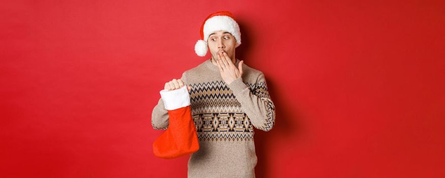 Concept of winter holidays, new year and celebration. Surprised adult man holding christmas stocking with presents inside, looking amazed at bag, standing over red background in santa hat.