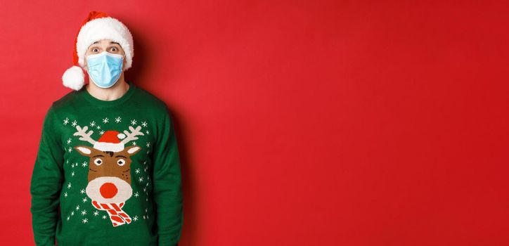 Concept of new year, covid-19 and social distancing. Surprised guy in santa hat, medical mask and christmas sweater looking amazed at camera, standing over red background.