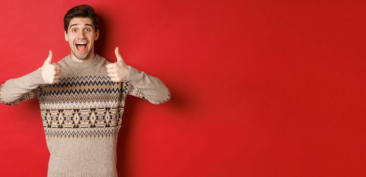 Portrait of cheerful man in christmas sweater like something good, showing thumbs-up and smiling amazed, praise new year party, standing over red background.