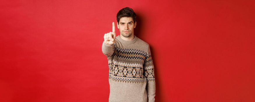 Image of serious handsome man extending one finger to stop you, telling no, give warning, standing over red background in winter sweater.