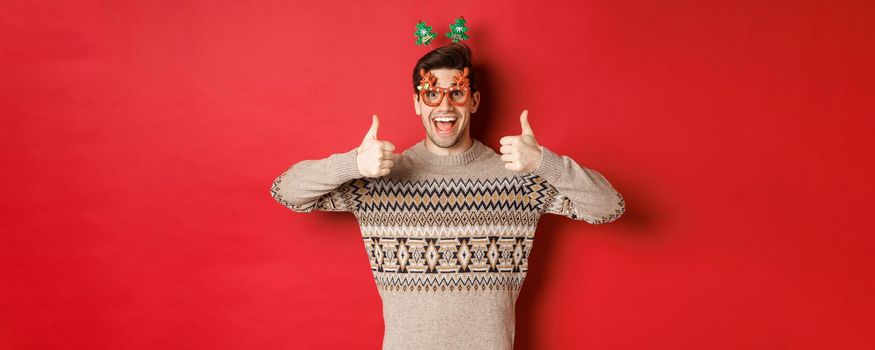Portrait of satisfied and happy man in christmas sweater and party glasses, showing thumbs-up, wishing happy new year, standing over red background.