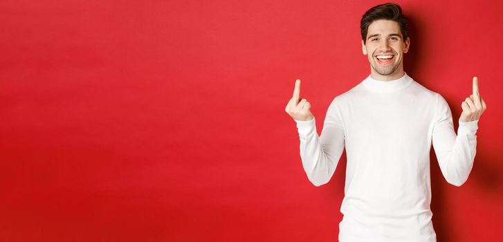 Image of rude and unbothered man laughing while showing middle-fingers, telling to fuck-off, standing over red background.