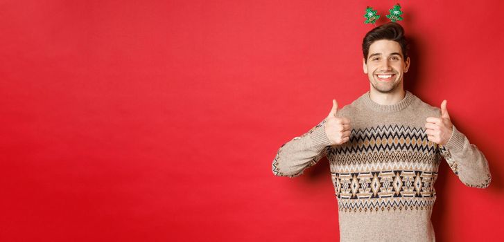 Concept of winter holidays, christmas and celebration. Cheerful bearded guy in sweater, showing thumbs-up in approval and smiling, enjoying new year party, red background.