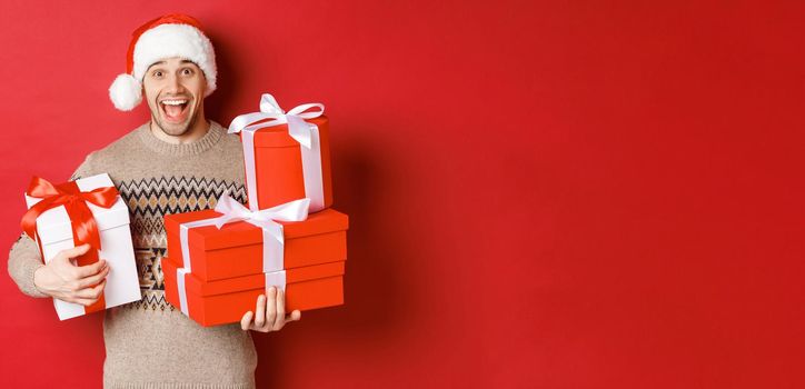 Concept of winter holidays, new year and celebration. Image of happy guy enjoying christmas, holding pile of presents and smiling amused, standing over red background.