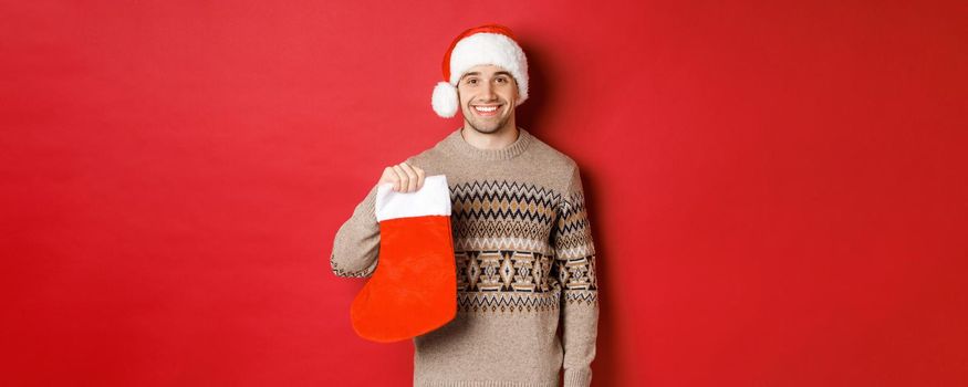 Concept of winter holidays, new year and celebration. Image of handsome smiling man in santa hat and sweater, holding christmas stocking for presents and candies, standing over red background.