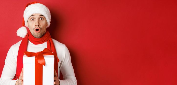 Concept of winter holidays, christmas and lifestyle. Close-up of surprised handsome guy in santa hat and scarf, looking amazed and holding new year gift, standing over red background.