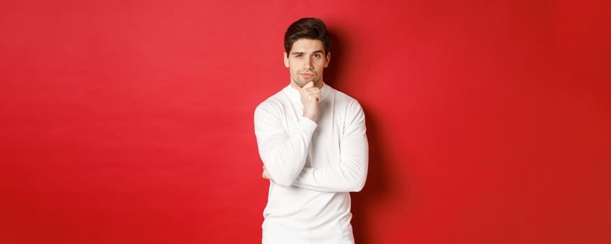 Image of thoughtful handsome man making assumption, thinking and looking at camera, standing in white sweater against red background.