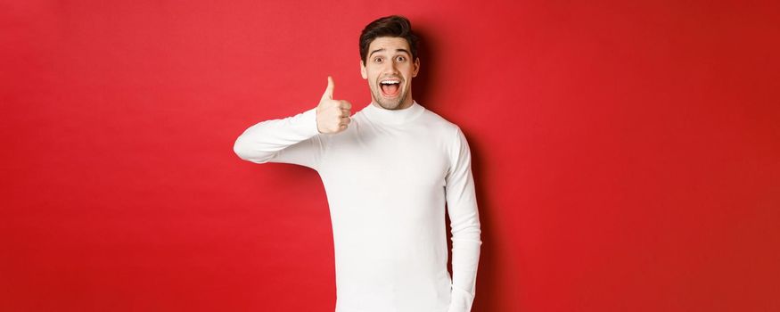 Concept of winter holidays, christmas and lifestyle. Excited good-looking man in white sweater, like good product, showing thumb-up in approval and smiling amazed, red background.