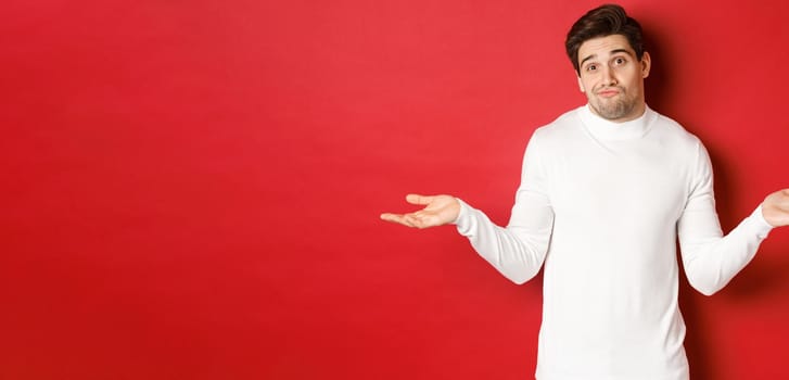 Portrait of clueless handsome guy, shrugging with hands spread sideways, being unaware, dont know anything, standing over red background.