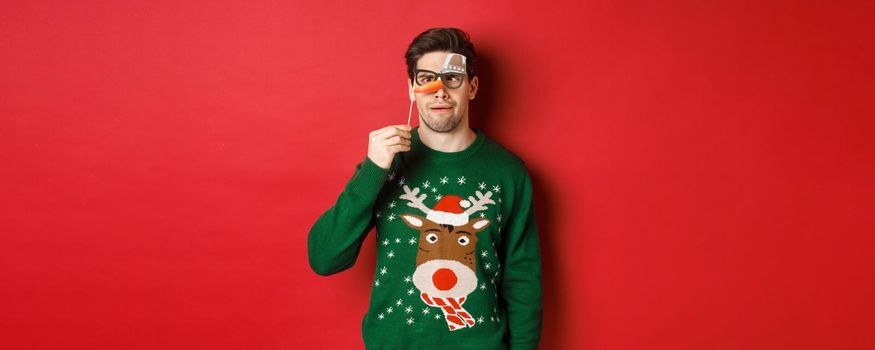 Funny man in christmas sweater and party mask, celebrating winter holidays, showing funny faces, standing over red background.