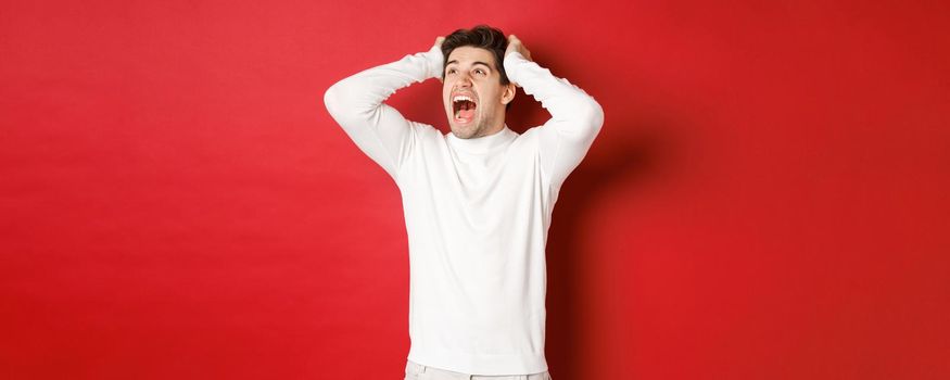 Portrait of frustrated man in white sweater, screaming in panic and looking at upper left corner, have terrible problem, standing over red background.