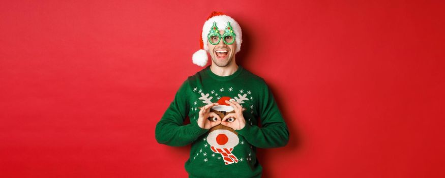 Portrait of carefree handsome man in santa hat and party glasses, making fun of his christmas sweater, looking happy over red background.