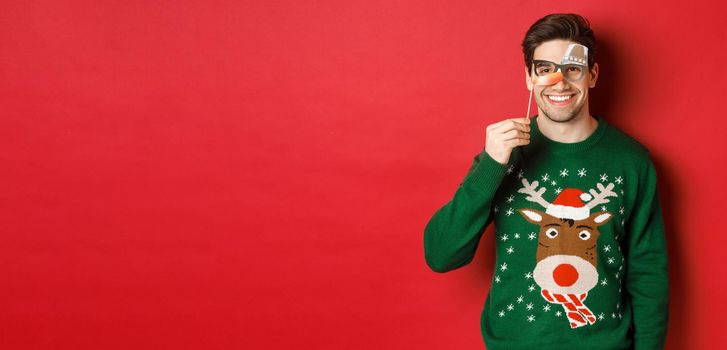 Funny man in christmas sweater and party mask, celebrating winter holidays, smiling happy, standing over red background.