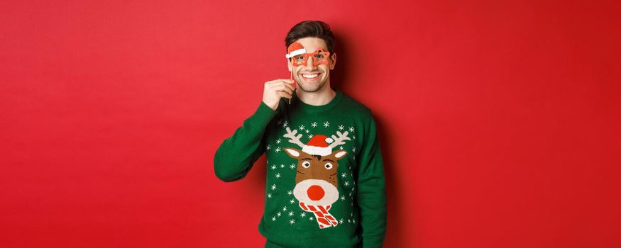 Portrait of handsome smiling man in christmas sweater and party glasses, celebrating new year and having fun, standing against red background.