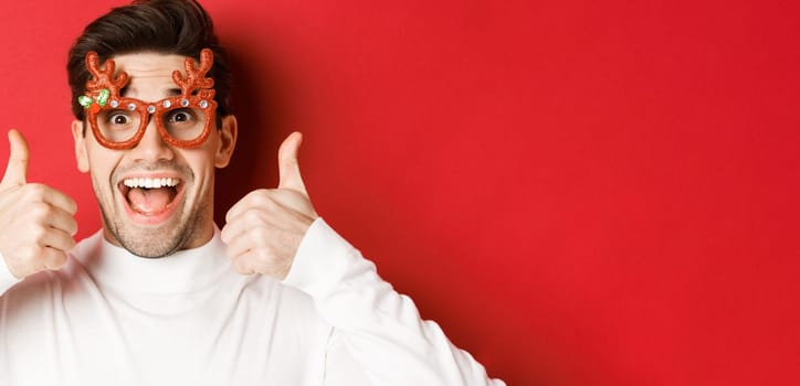 Concept of winter holidays, christmas and celebration. Close-up of excited handsome man in party glasses, smiling and showing thumbs-up in approval, standing over red background.