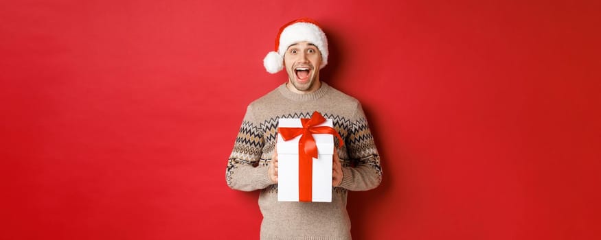 Image of excited handsome man receiving christmas gift, wearing santa hat and winter sweater, shouting for joy, holding present and standing over red background.