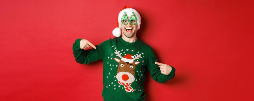 Image of happy man in party glasses and santa hat, pointing at his christmas sweater and smiling, standing over red background.