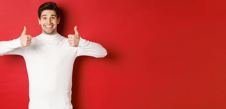 Cheerful good-looking male model in white sweater, showing thumbs-up in approval, like something good, standing over red background and smiling pleased.