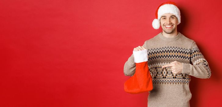 Concept of winter holidays, new year and celebration. Handsome smiling man prepared gifts for kids, pointing at christmas stocking bag, standing over red background.