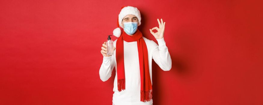 Concept of covid-19, christmas and holidays during pandemic. Cheerful man celebrating new year in medical mask and santa hat, recommending hand sanitizer, showing okay sign and winking.