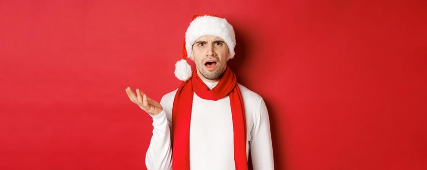 Concept of christmas, winter holidays and celebration. Portrait of confused man in santa hat and scarf, frowning and looking perplexed, standing over red background.