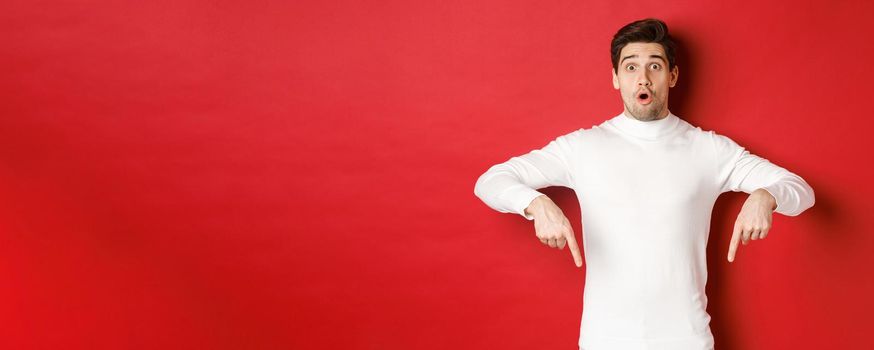 Image of surprised attractive guy in white sweater, showing announcement, pointing fingers down and looking amazed, standing over red background.