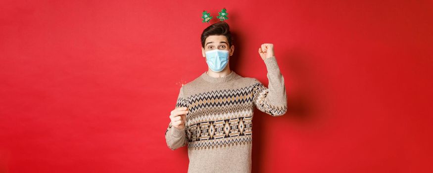 Concept of new year, covid-19 and social distancing. Happy man celebrating in medical mask, holding sparkler and enjoying party, standing over red background.