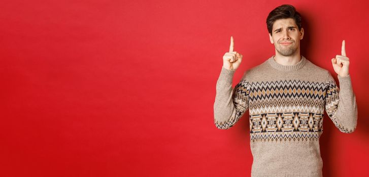 Image of skeptical and unamused handsome guy, showing bad christmas offer, pointing fingers up and grimacing displeased, express dislike, standing over red background.