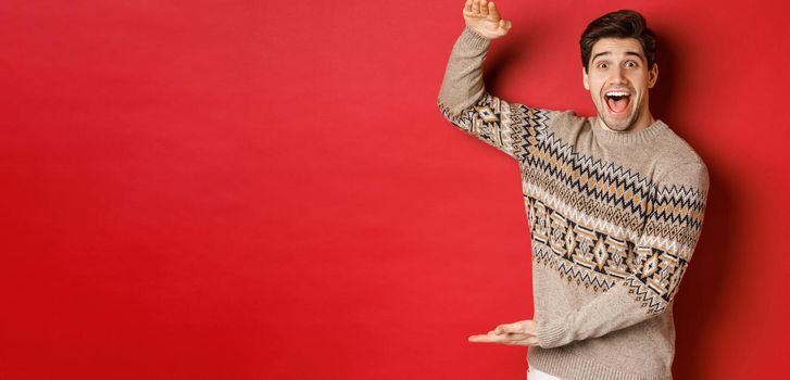 Portrait of excited and happy handsome man in christmas sweater, showing something big, holding large gift for holidays, standing over red background.