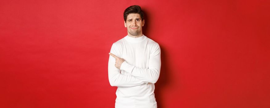 Concept of winter holidays, christmas and lifestyle. Image of skeptical young man in white sweater, dont like something, pointing finger left and looking with dismay, standing over red background.