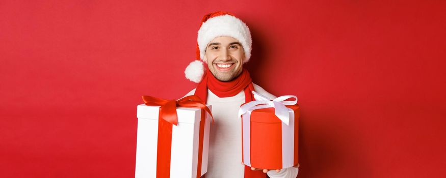 Concept of winter holidays, christmas and lifestyle. Close-up of smiling handsome guy in santa hat and scarf, wishing happy new year and holding gifts, standing with presents over red background.