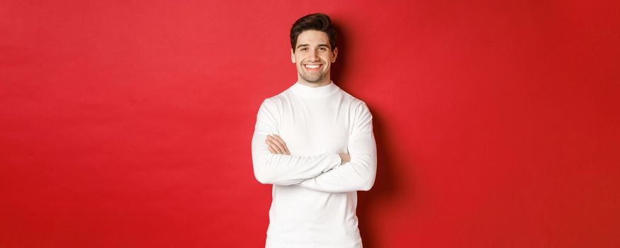 Concept of winter holidays, christmas and lifestyle. Portrait of confident good-looking man with bristle, wearing white sweater, cross arms on chest and smiling satisfied.