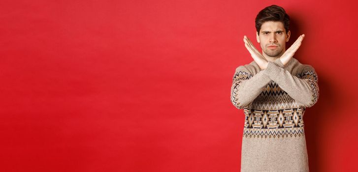 Image of angry and serious handsome man in christmas sweater, telling no or stop, showing cross gesture to restrict you from something, prohibit action, standing over red background.