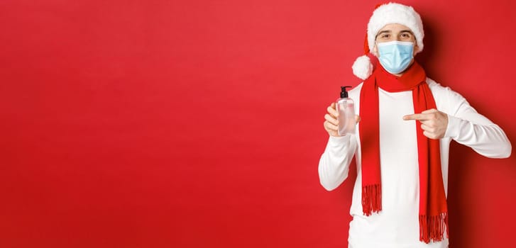 Concept of covid-19, christmas and holidays during pandemic. Handsome happy man in santa hat and medical mask, recommending use hand sanitizer, standing over red background.