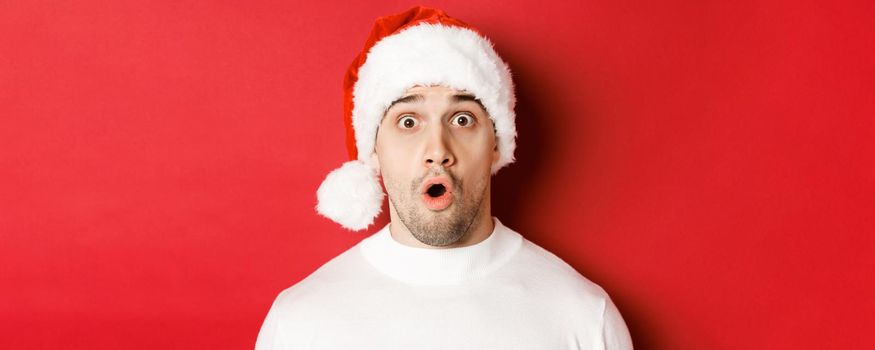 Close-up of attractive surprised man in santa hat, saying wow and looking at something interesting, standing against red background.