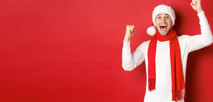 Image of joyful caucasian man in santa hat and scarf, shouting for joy and raising hands, celebrating victory or win, triumphing over red background.