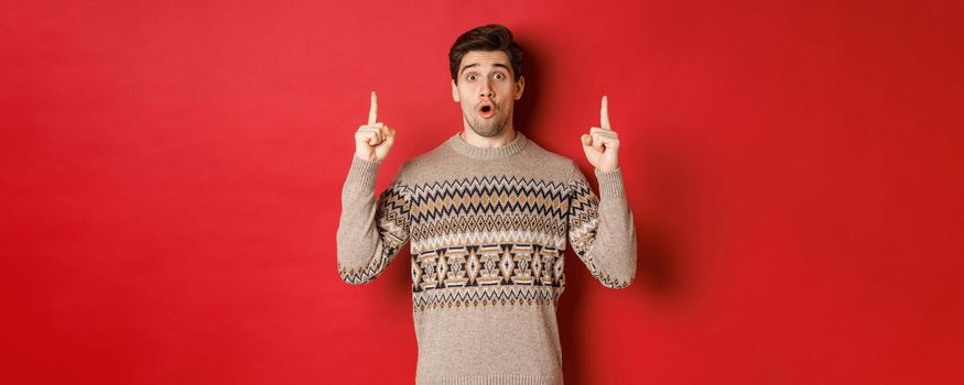 Portrait of surprised and amazed handsome guy showing christmas promo offer, wearing xmas sweater and standing over red background.