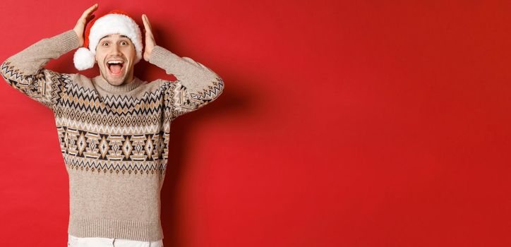 Image of surprised and happy young man in santa hat and christmas sweater, receiving amazing gift, standing excited against red background.
