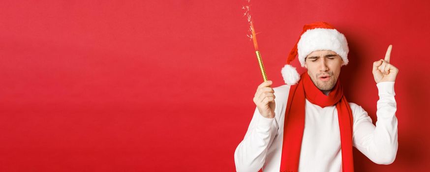 Concept of christmas, winter holidays and celebration. Attractive guy enjoying new year party, dancing with sparkler, wearing santa hat and scarf, standing over red background.