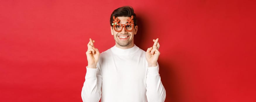 Close-up of excited handsome man, wearing party glasses and white sweater, crossing fingers for good luck, celebrating christmas and waiting wish, anticipating gifts, standing over red background.