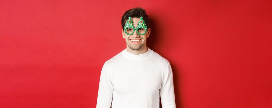 Concept of christmas, winter holidays and celebration. Close-up of handsome man in white sweater and party glasses, enjoying new year, standing over red background.