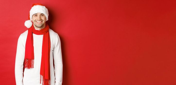 Happy caucasian guy celebrating christmas and smiling, wearing santa hat, scarf and white sweater, enjoying new year holidays, standing over red background.