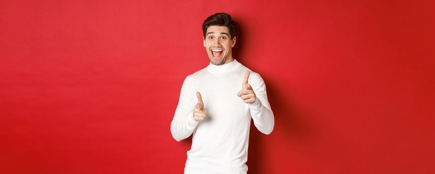 Concept of winter holidays, christmas and lifestyle. Cheerful handsome guy in white sweater congratulating you, pointing fingers at camera and wishing happy new year, standing over red background.