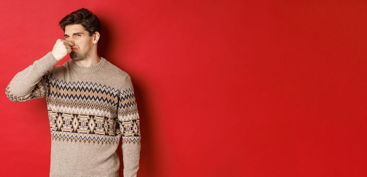 Image of disgusted young man in winter sweater, shut nose and grimacing from bad smell, standing over red background.