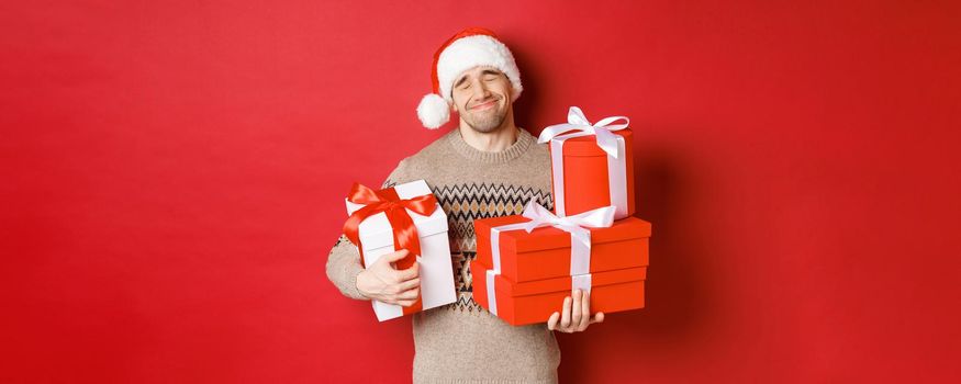 Concept of winter holidays, new year and celebration. Portrait of lovely smiling man receiving pile of presents, holding gifts and being touched with surprise, standing over red background grateful.