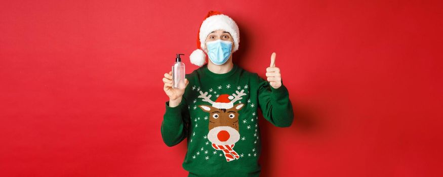Concept of new year, coronavirus and social distancing. Portrait of cheerful man in santa hat, christmas sweater and medical mask, showing thumb-up and recommending hand sanitizer.