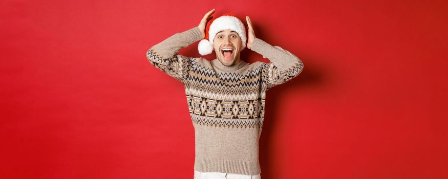 Image of surprised and happy young man in santa hat and christmas sweater, receiving amazing gift, standing excited against red background.