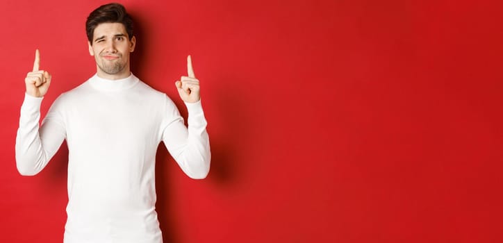 Concept of winter holidays. Disappointed handsome man do not recommend promo, pointing fingers up and grimacing displeased, standing over red background in white sweater.