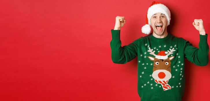 Portrait of cheerful attractive man celebrating new year, wearing green sweater and santa hat, shouting for joy, winning or triumphing, standing over red background.